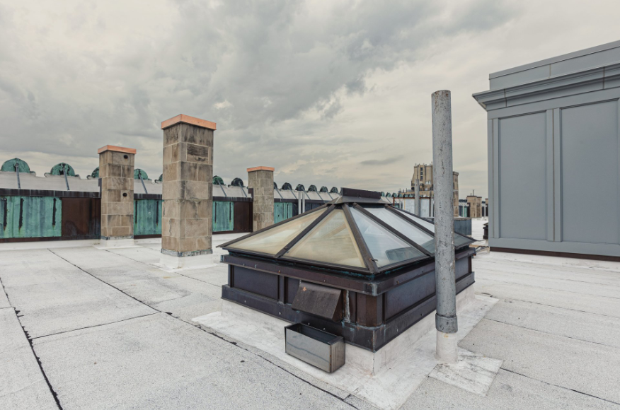 Flat commercial rooftop with a pyramid skylight, vent pipe, and chimney structures under a cloudy sky.