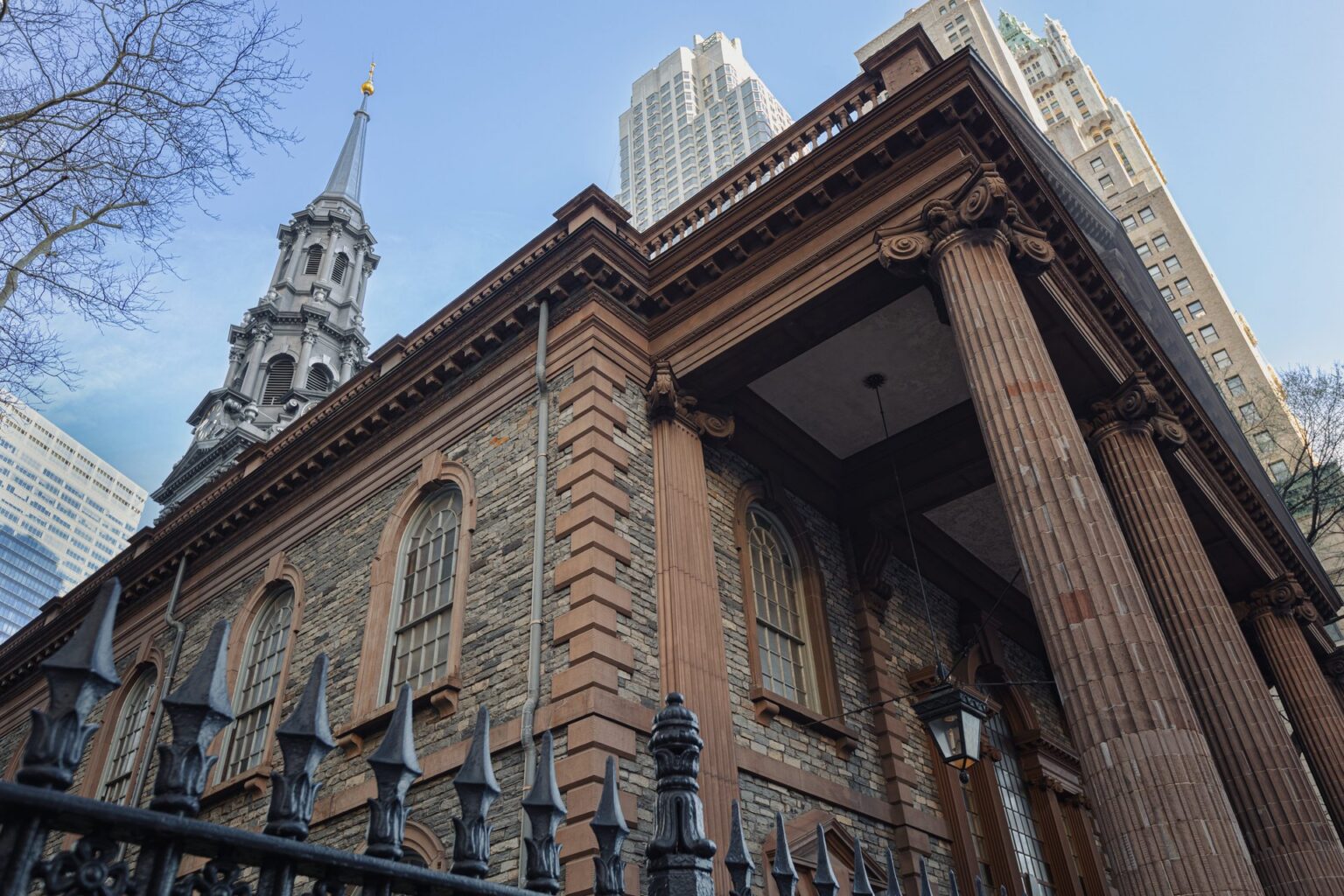 Historic stone church facade with tall columns and steeple surrounded by modern high-rise buildings.
